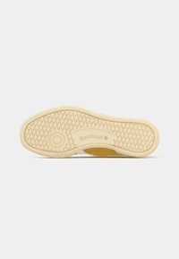 Reebok tenisica s bež gumom na potplatu, s teksturiranim uzorkom i kružnim logom. Ima glatku završnu obradu i blage utore za prianjanje.