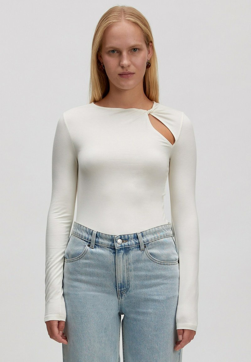 Weißer Langarmbodysuit mit rundem Ausschnitt und einem Ausschnitt an der Schulter, kombiniert mit hellblauen hochgeschnittenen Jeans.