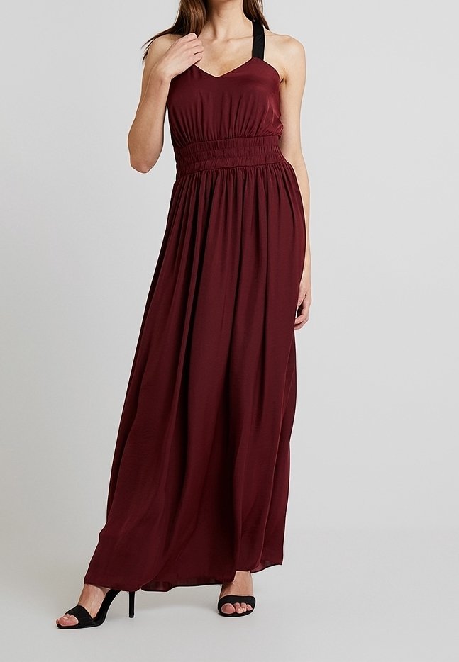Femme portant une robe longue bordeaux sans manches avec des bretelles noires et des sandales noires à talons hauts, debout devant un fond gris uni.