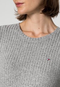 Pull gris côtelé en tissu texturé ; doté d'un col rond et d'un petit logo en rouge, blanc et bleu sur le côté gauche.