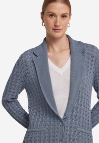 Ljusblå stickad cardigan med ett texturerat mönster, stor sjalkrage, två knappar och framfickor. Lager på en vit topp.