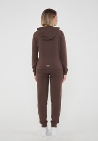 Tuta con cappuccio marrone realizzata in morbido tessuto, caratterizzata da un design aderente e polsini a coste. Logo dorato distintivo sul retro. Sneakers bianche.