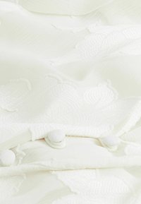Gros plan sur un tissu blanc texturé avec une broderie florale en relief et trois boutons recouverts de tissu le long d'un bord plié.