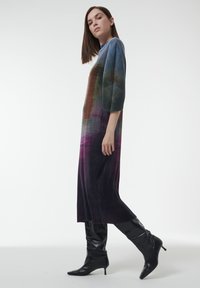 Lang, aansluitend jurk van meerkleurige geribbelde stof, met een verloop van blauw naar paars. Draagt een paar zwart, aansluitend laarzen tot de knie.