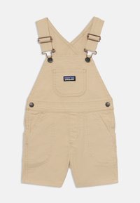 Salopette courte en coton beige avec bretelles réglables, poche poitrine avant et deux poches latérales. Comprend des accessoires en métal et des accents cousus.