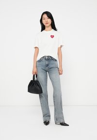 Camiseta blanca con gráfico de corazón rojo, jeans acampanados azules, bolso negro de tiras con detalles recogidos y zapatos negros de punta.