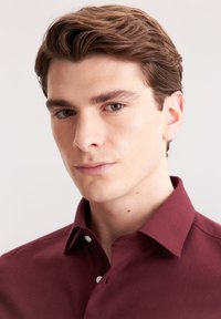 Camicia bordeaux in un tessuto morbido con colletto strutturato, caratterizzata da bottoni bianchi e un sottile dettaglio di cucitura sopra il bottone sinistro.
