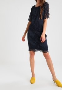 Robe en dentelle bleu marine avec manches courtes et silhouette ajustée. Présente des motifs floraux complexes et un ourlet festonné. Associée à des mules jaunes.