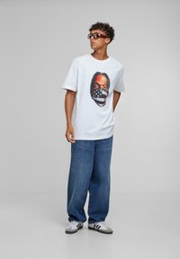 White short-sleeve t-shirt featuring a graphic of a masked face. Worn with loose blue jeans and gray sneakers with black stripes.