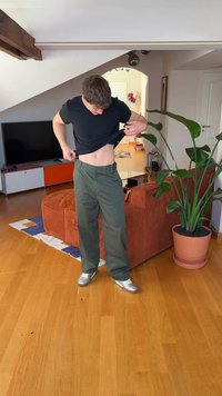 Cămașă neagră cu mâneci scurte, pantaloni largi verde măsliniu și pantofi argintii. Cadru interior cu o plantă și o canapea maro în fundal.