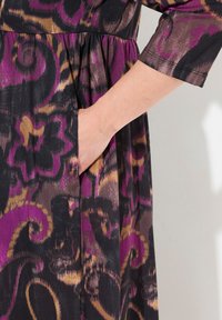 Robe en tissu noir avec des motifs tourbillonnants audacieux en violet, marron et or. Comprend une poche latérale avec des détails de taille froncée.