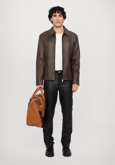 Bruine leren ritsjack, witte shirt, zwarte broek en zwarte gesp laarzen. Oranje tas met handvatten en schouderband. Gladde textuur.