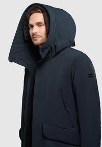Giacca imbottita blu scuro con un grande cappuccio, chiusura con zip e tasche laterali. Presenta un logo sul braccio e una texture liscia.