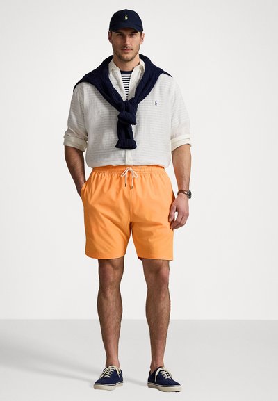 Pantaloni scurți de baie portocalii cu talie elastică, purtați cu o cămașă albă cu nasturi și un pulover bleumarin legat în jurul umerilor. Capp bleumarin și pantofi.