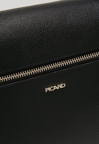 Surface en cuir noir texturé avec une fermeture éclair dorée horizontale et le nom de marque doré "PICARD" sous la fermeture éclair.