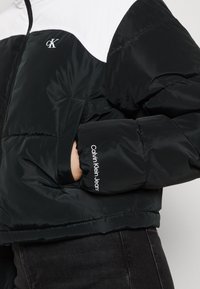 Giubbotto pom pom nero con pannello superiore bianco, chiusura a zip e logo ricamato "Calvin Klein Jeans" sul polsino. Materiale liscio e lucido.