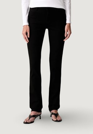 Persona vistiendo pantalones ajustados negros, camiseta blanca de manga larga y sandalias negras de tacón con punta abierta, de pie sobre un fondo claro.