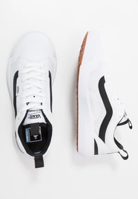White sneakers with black accents, textured mesh upper, white rubber sole with grip, featuring a pull tab and lacing system. Brand logo present.