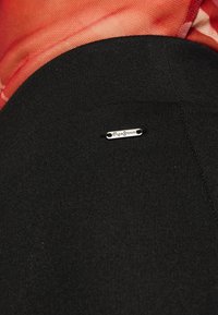 Zwarte stof met een klein zilveren label met de tekst "Pepe Jeans" nabij de naad, in contrast met de rode gestructureerde stof erboven.