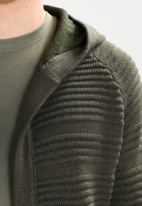 Olivgrön stickad cardigan med huva, med ribbad struktur och släta detaljer längs kragen, lager på lager över en matchande t-shirt.