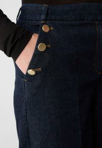 Jeansy z ciemnoniebieskiego denimu z widokiem z bliska na trzy metalowe guziki po boku, z pomarańczowymi przeszyciami i gładką fakturą.