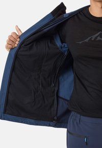 Person iført sort skjorte og blå bukser holder en åben blå jakke for at vise det sorte netfor og den indvendige lynlåslomme.