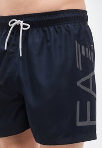 Μπλε ναυτικές βερμούδες κολύμβησης από ελαφρύ υλικό, με κορδόνι στη μέση και τολμηρό, διάτρητο σχέδιο στο πλάι.