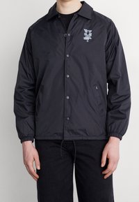 Mustat nyloni takki, jossa on painonappikiinnitys, elastiset hihansuut ja kiristysnyöri helmassa. Mukana on sävy sävyyn -logo vasemmassa rinnassa ja kaksi sivutaskua.