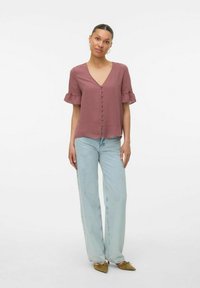 Blusa a maniche corte color rosa polveroso con scollatura a V, bottoni rivestiti in tessuto e maniche arricciate, abbinata a jeans a gamba larga blu chiaro e scarpe verdi oliva.