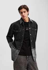 Sort denimjakke med en gradient overgange til grå, der har to brystlommer, trykknapper og en krave over en sort t-shirt.
