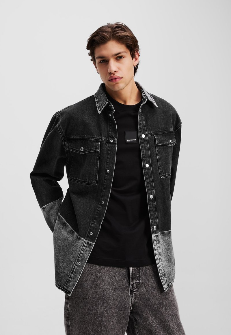 Sort denimjakke med en gradient overgange til grå, der har to brystlommer, trykknapper og en krave over en sort t-shirt.
