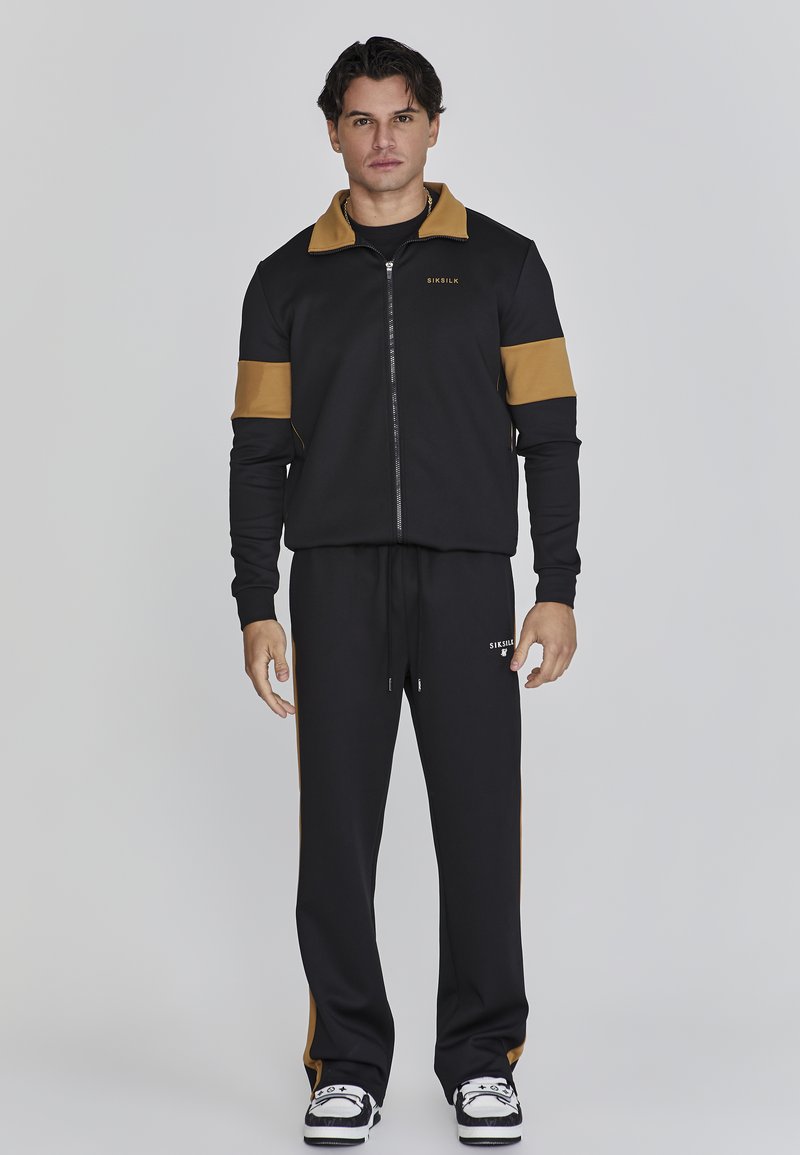 Veste zippée noire avec des rayures brunes et un logo, assortie de pantalons noirs avec des rayures sur les côtés et un cordon de serrage. Fabrication en tissu lisse.