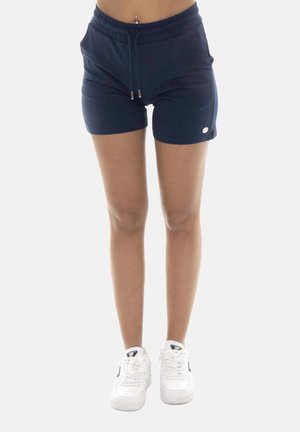 Persona che indossa pantaloncini blu navy con coulisse e scarpe da ginnastica bianche, in piedi contro uno sfondo chiaro e uniforme.