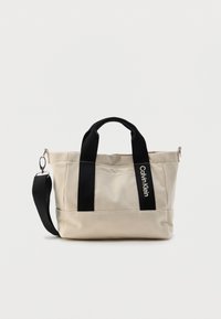 Μπεζ τσάντα χειρός από καμβά με μαύρες λαβές και λουρί ώμου. Διαθέτει ραμμένες λεπτομέρειες και λογότυπο στο πλάι. Συμπαγής σχεδίαση.