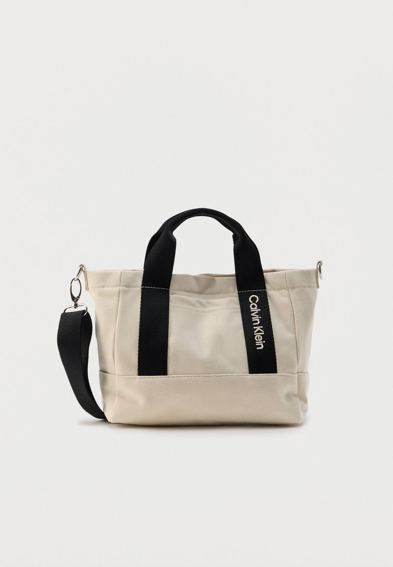 Beežist lõuendist kott mustade käepidemete ja õlarihmaga. Omab õmblusdetailid ja logo külgmisel küljel. Kompaktne disain.