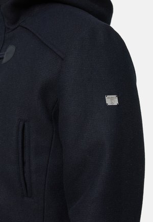 Marineblå hettejakke laget av teksturert stoff, med toveis glidelås, sidelommer og en liten metallisk logo på ermet.