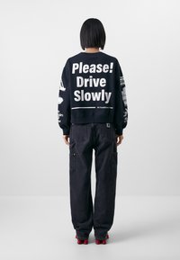 Felpa nera con testo bianco "Per favore! Guida piano" sulla schiena; abbinata a pantaloni cargo neri larghi; scarpe rosse visibili.