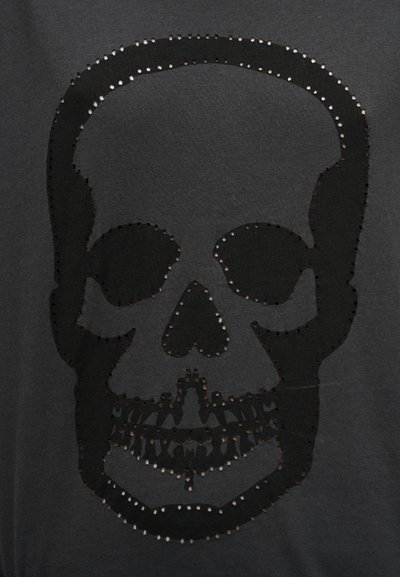 Μαύρο μπλουζάκι με μεγάλο σχέδιο κρανίου από κοψίματα περιβαλλόμενα από μικρές ασημένιες μεταλλικές λεπτομέρειες, δημιουργώντας ένα υφασμένο εφέ.