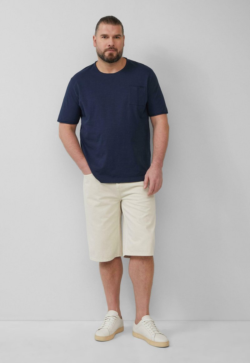 Navy katoen T-shirt met een borstzak, crèmekleurige shorts en witte sneakers met lichte zolen. Eenvoudige, casual outfit op een model.