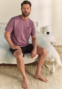 Levander színű, rövid ujjú póló elülső zsebekkel, fekete sorttal párosítva. A modell egy ágyon ül, amelyen egy textúrált krém színű szőnyeg található.