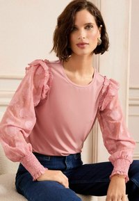 Top rosa con maniche corte trasparenti e svolazzanti con motivi floreali; corpo in tessuto liscio, abbinato a jeans in denim scuro aderenti.