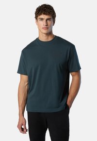 Uomo che indossa una t-shirt a maniche corte di colore verde scuro con un piccolo logo "North Sails" e pantaloni neri, mentre posa su uno sfondo chiaro e semplice.