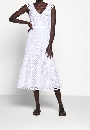 Femme portant une robe midi blanche sans manches en broderie anglaise avec un col à volants et des sandales slides noires, debout devant un fond uni.
