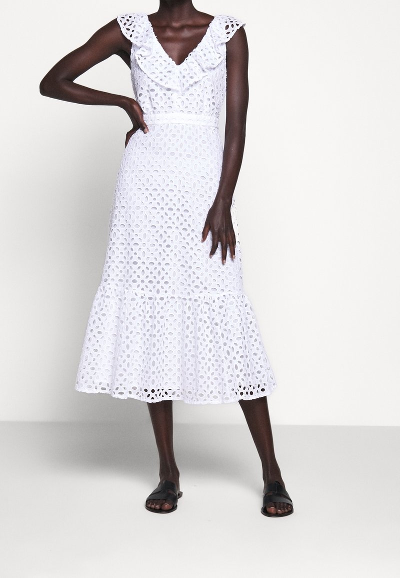 Femme portant une robe midi blanche sans manches en broderie anglaise avec un col à volants et des sandales slides noires, debout devant un fond uni.