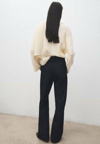Femme aux longs cheveux foncés portant un pull côtelé crème et un pantalon large taille haute noir, debout contre un mur blanc.