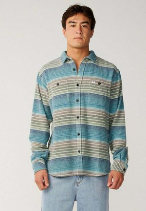 Joven con cabello corto y oscuro que lleva una camisa de botones a rayas azul, verde y beige y vaqueros azul claro, de pie frente a un fondo liso.