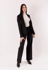 Giacca da completo nera sopra un dolcevita bianco a coste, abbinata a pantaloni neri a gamba larga e décolleté nere con punta a punta. Design semplice e su misura.