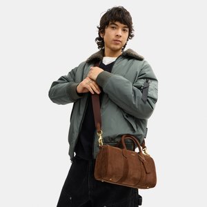 Ung person iført grøn bomberjakke og sorte bukser, der holder en brun ruskindstaske med skulderrem mod en hvid baggrund.