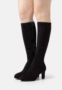 Botas altas de tacón bajo negras, fabricadas en suave ante, con puntera redonda, detalle de cremallera vertical y un tacón rectangular delgado. Textura lisa sin patrones.
