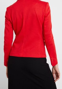 Blazer rouge à coupe ajustée, tissu lisse, manches longues et trois boutons décoratifs sur chaque manche, porté sur une jupe noire côtelée.